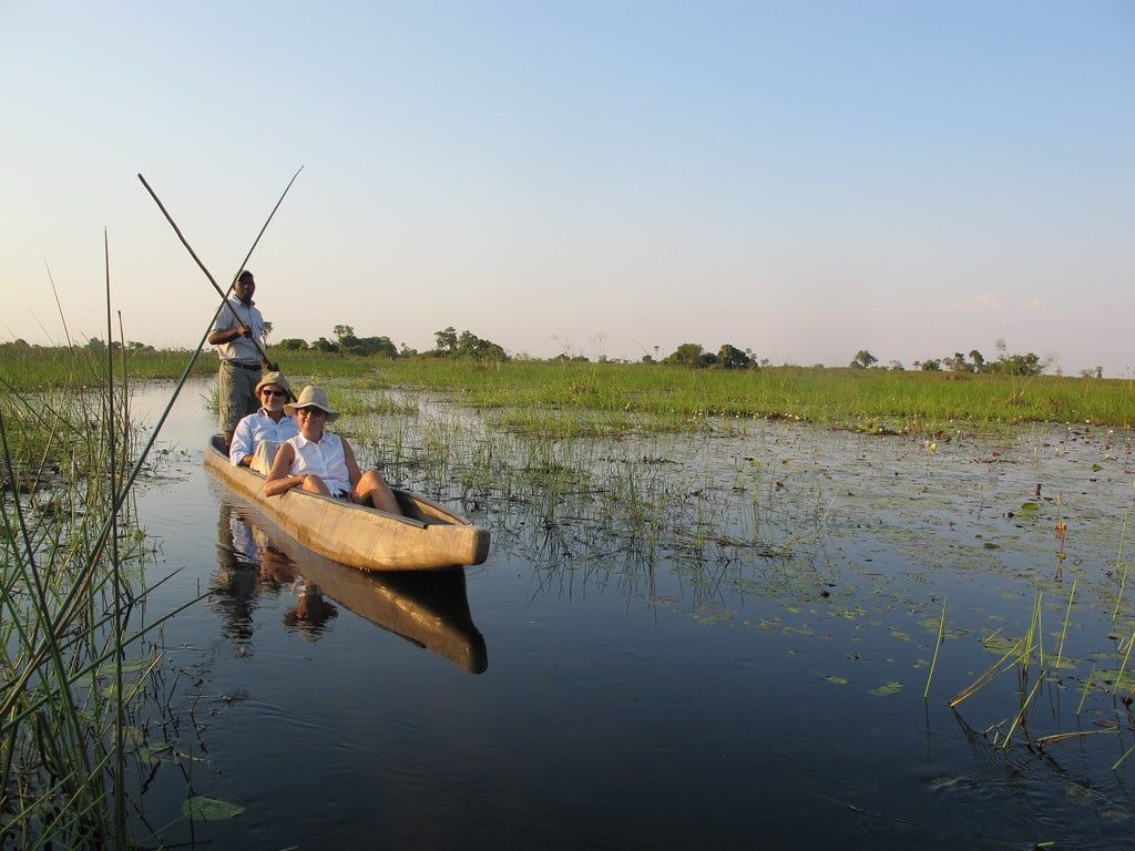 Ein Guide stakt Touristen in einem traditionellen Mokoro-Kanu durch einen flachen, von Schilf gesäumten Kanal im Okavango-Delta.
